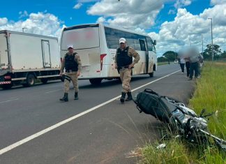 Acidente entre moto e transporte coletivo termina em morte em Uberlândia Local do acidente isolado com fita zebrada, mostrando a motocicleta caída no asfalto próxima a um ônibus urbano e policiais militares ao fundo