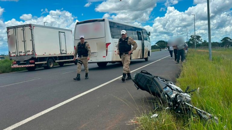 Local do acidente isolado com fita zebrada, mostrando a motocicleta caída no asfalto próxima a um ônibus urbano e policiais militares ao fundo