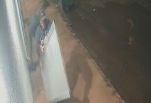 Até a porta: Ação de criminosos surpreende moradores do bairro Morumbi Imagem de câmera de segurança mostrando um homem de casaco escuro carregando uma porta de alumínio branca em uma rua à noite.