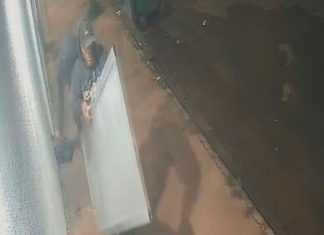 Até a porta: Ação de criminosos surpreende moradores do bairro Morumbi Imagem de câmera de segurança mostrando um homem de casaco escuro carregando uma porta de alumínio branca em uma rua à noite.