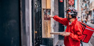 Atenção, Uberlândia: Prédios e condomínios verticais têm novas normas de entrega Entregador de aplicativo com mochila térmica entregando encomenda para porteiro na entrada de um prédio.